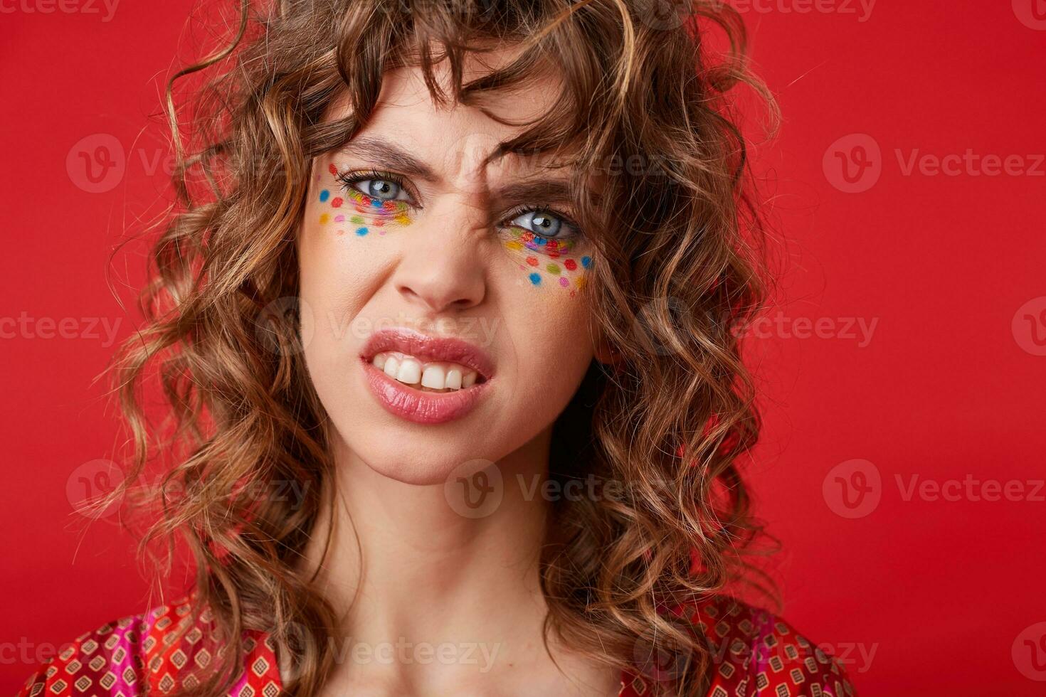 Studio shot of young brunette blue-eyed female with festive hairstyle looking at camera with pout and demonstrating her teeth while standing over red background photo