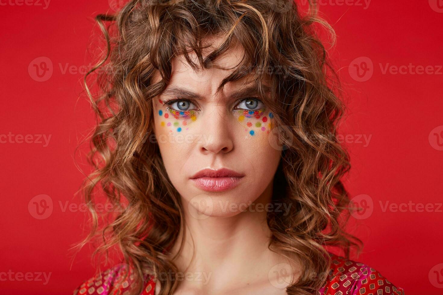 Severe pretty young curly woman in colored patterened top wearing festive makeup, frowning with folded lips, looking seriously to camera while isolated over red background photo
