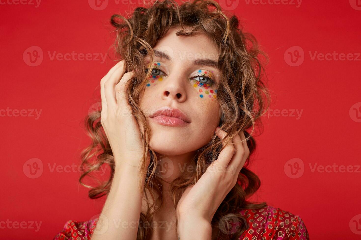 Indoor shot of lovely young lady with romantic hairstyle wearing motley patterned top, looking at camera with folded lips and keeping palms on her head, posing over red background photo
