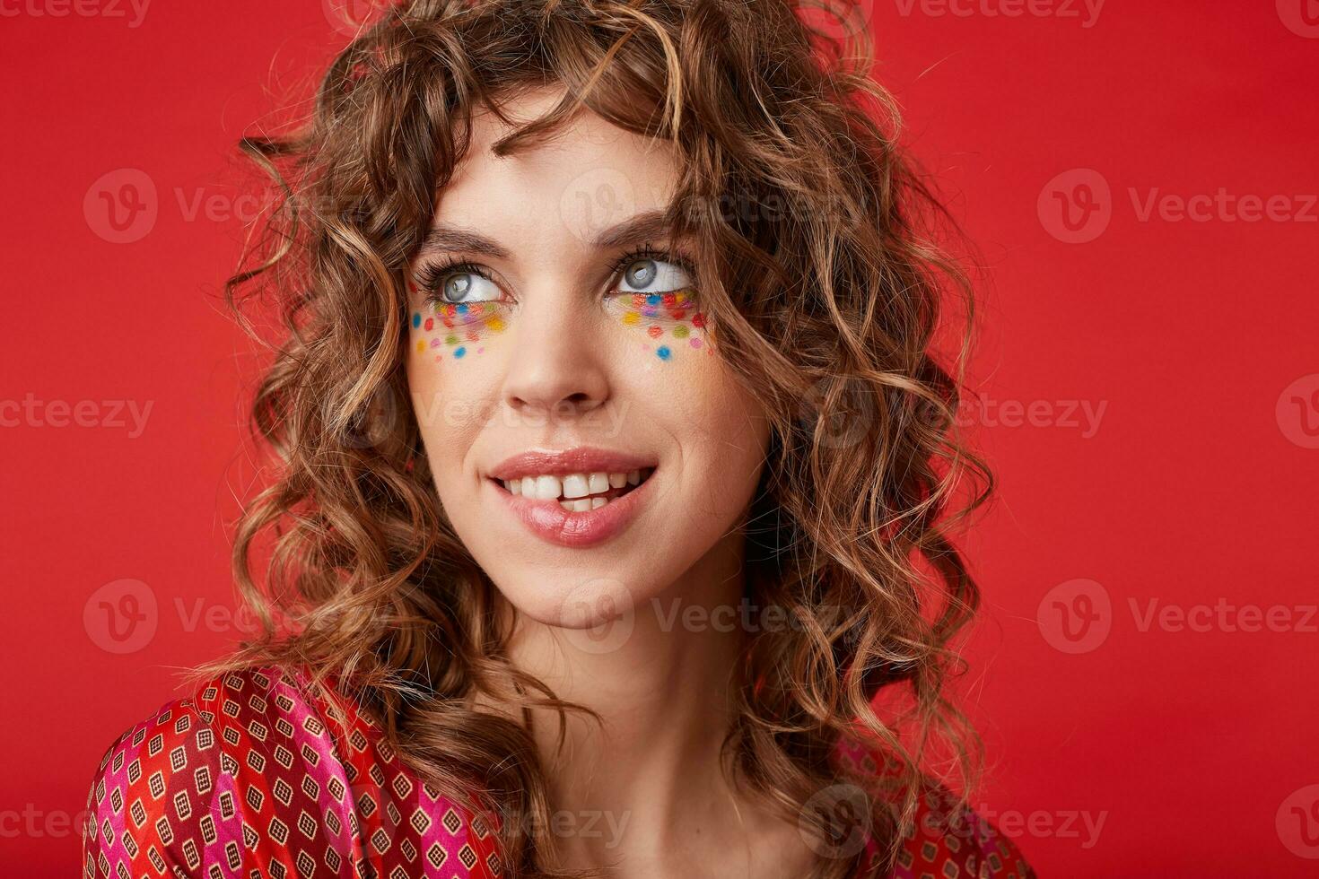 Beautiful young blue-eyed female with curly hair and with festive makeup looking aside with gentle smile and biting underlip, standing over red background in motley patterned top photo