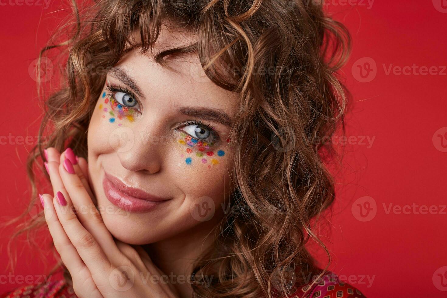 Positive pretty young female with romantic hairstyle and multicolored dots on her face looking at camera with tender smile and leaning chin on folded hands, standing over red background photo