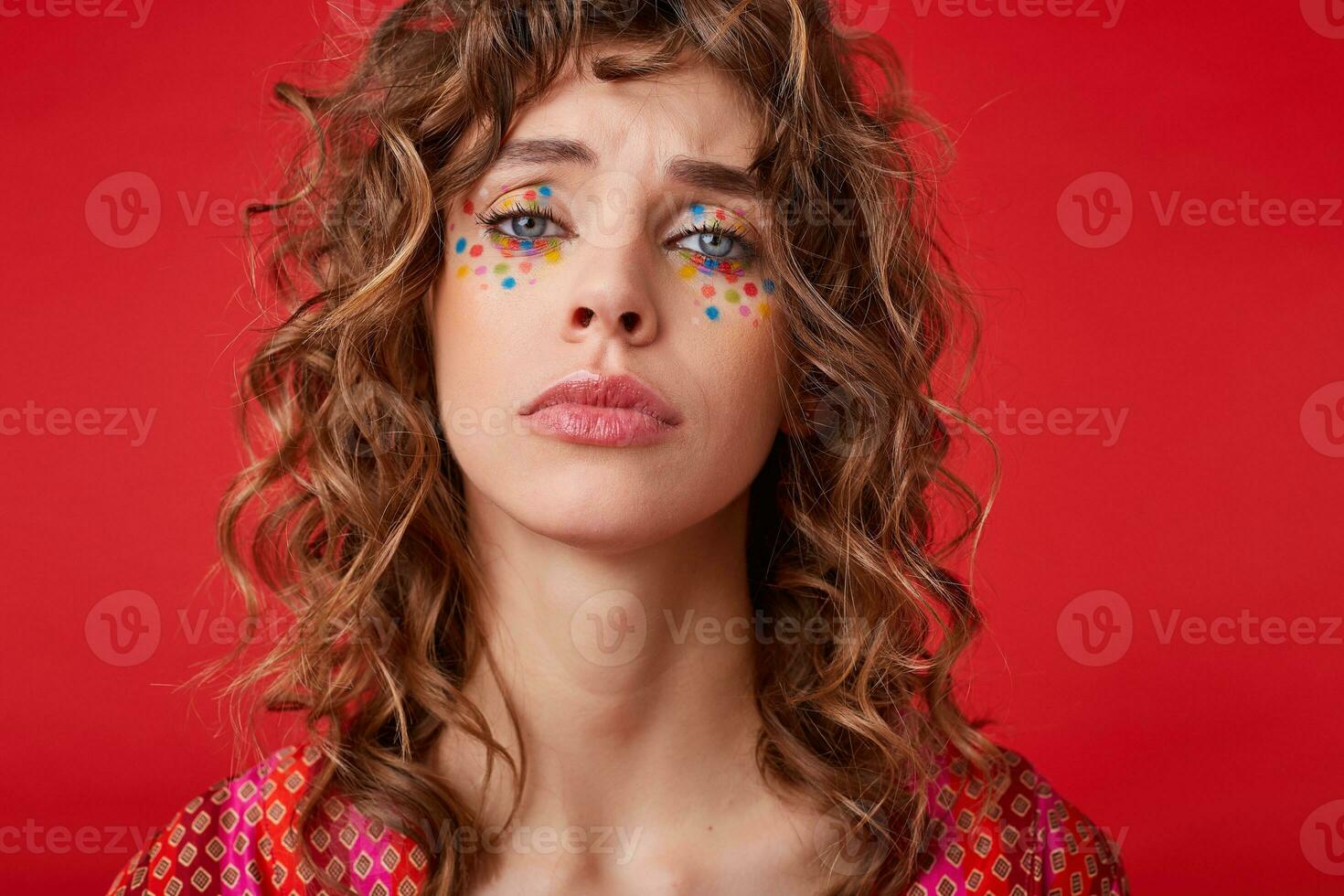 Upset young curly female with festive makeup wearing motley patterned top, frowning and looking to camera with folded lips, posing over red background photo