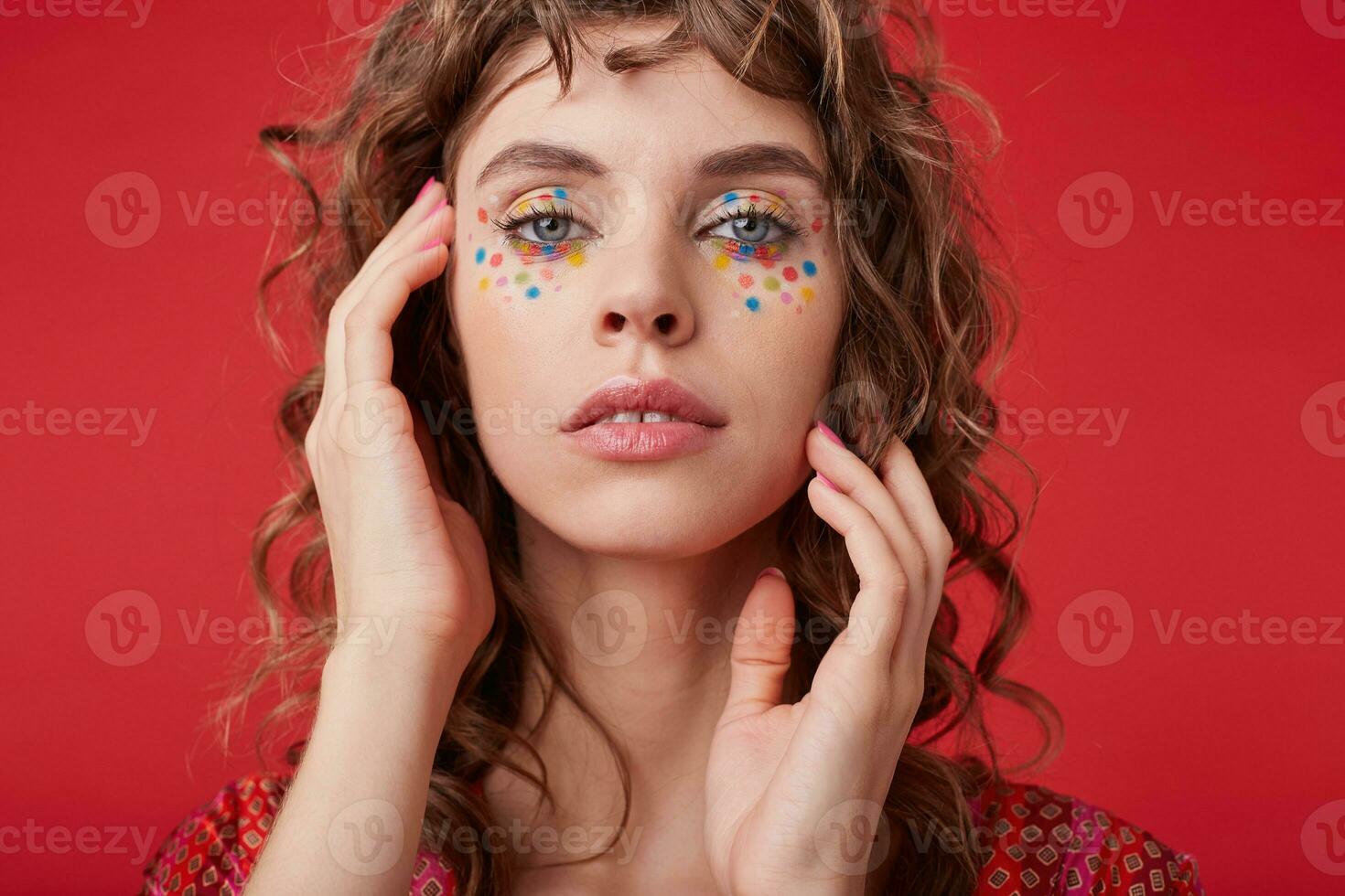 Beautiful blue-eyed curly woman with multicolored dots on her face wearing motley patterned top, keeping hands gently near her face, isolated over red background photo