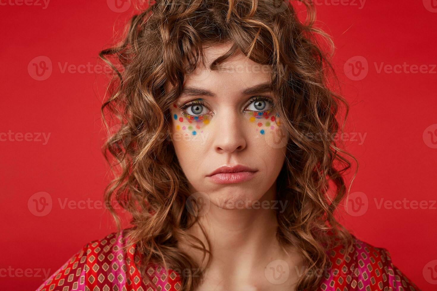 Studio photo of sad blue-eyed young woman with romantic hairstyle and festive makeup looking to camera wailfully, posing over red background in motley patterned top