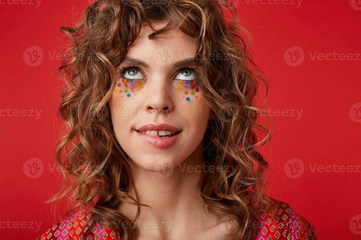 Portrait of young attractive curly woman with multicolored dots on her face looking upwards thoughtfully and biting underlip, wearing motley patterned top while posing over red background photo