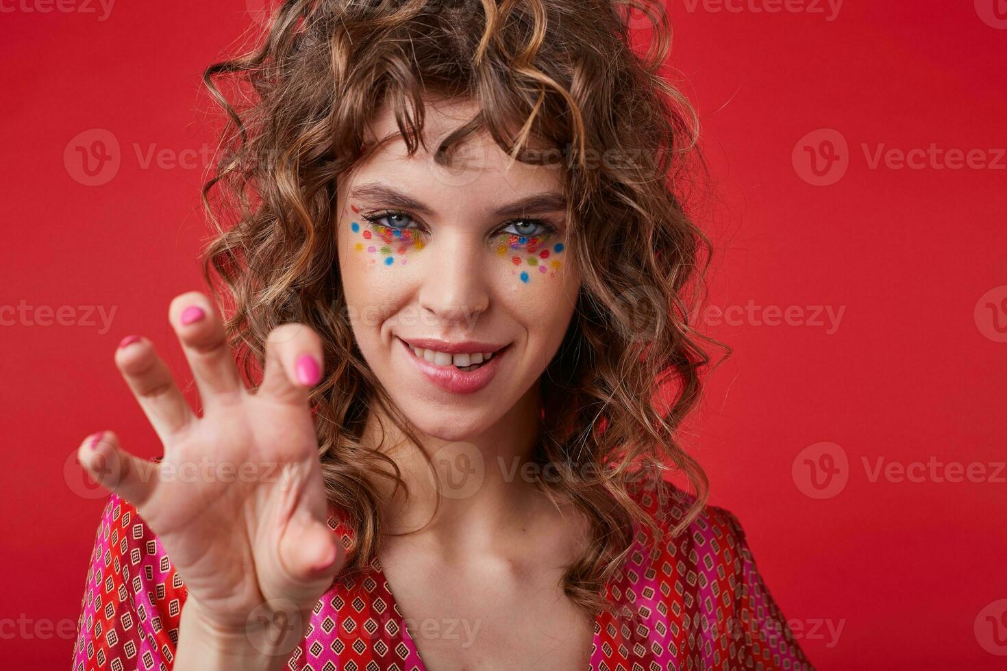 Coquettish young curly woman with multicolored dots on her face looking at camera and biting underlip, raising hand playfully while standing over red background in motley patterned top looking camera photo