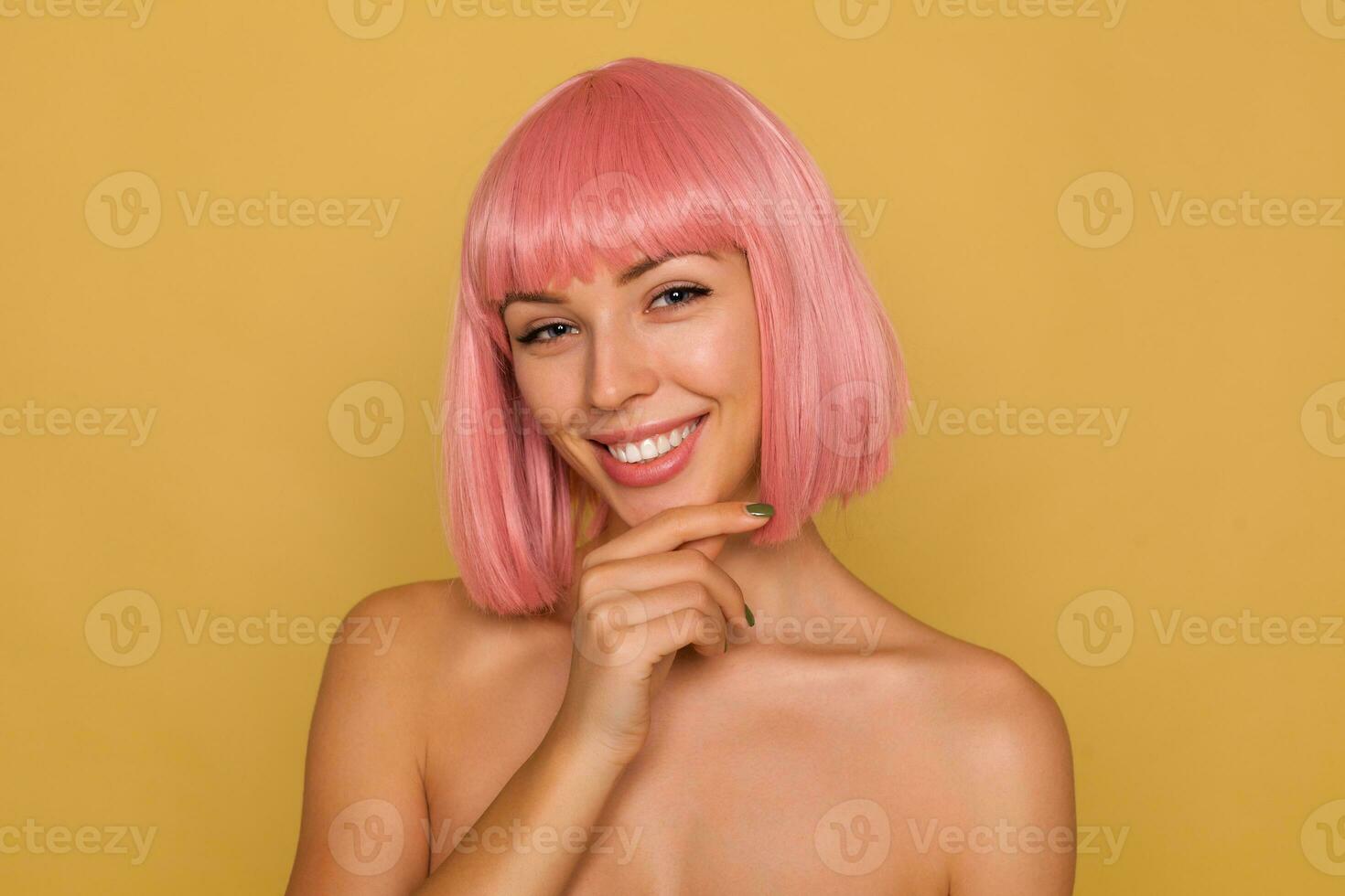 Portrait of young happy blue-eyed pink haired female looking happily at camera with charming smile, holding raised hand on her chin while posing over mustard background photo