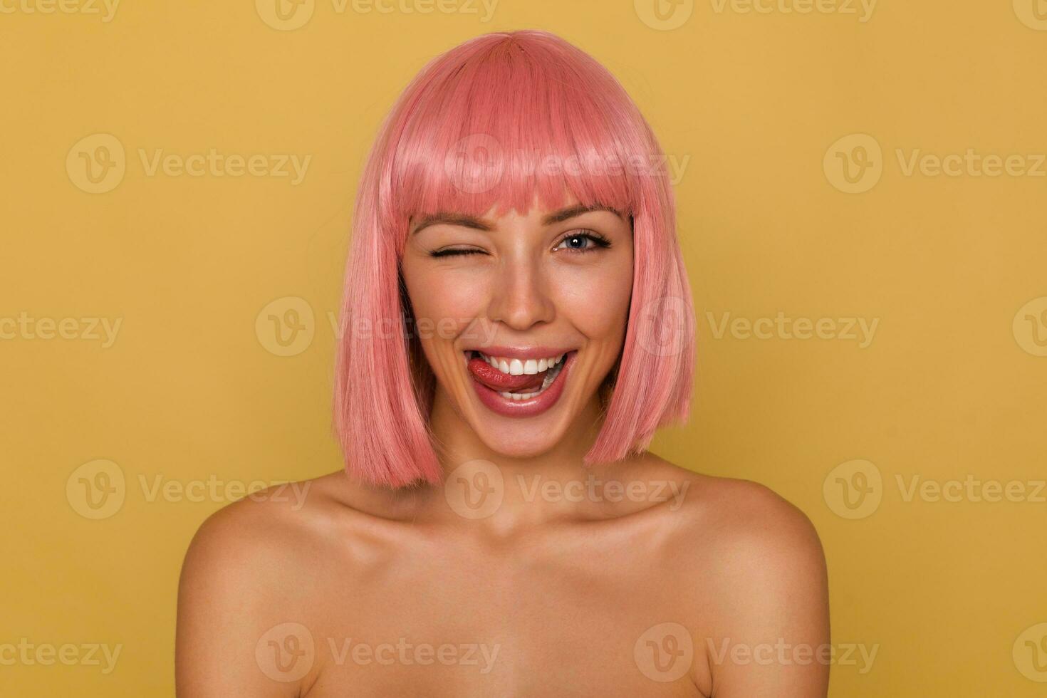 Coquettish young pretty cheerful pink haired female with bob hairstyle looking joyfully at camera and sticking out her tongue, winking happily at camera while standing over mustard background photo