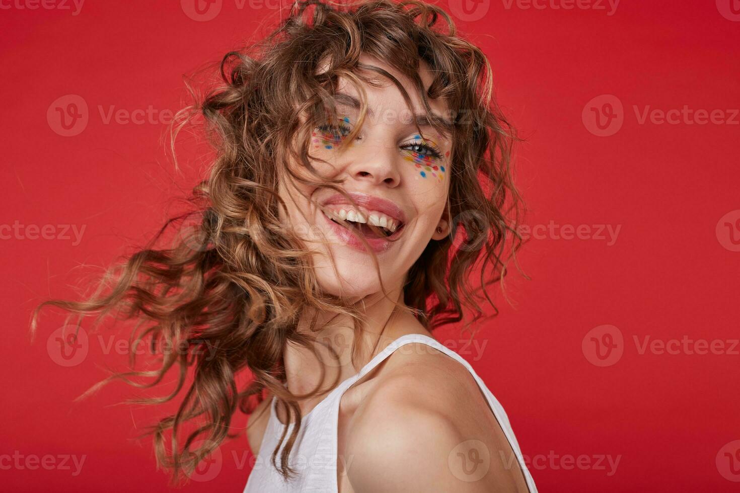 Studio photo of charming cheerful young woman with curly hair twisting her head, wearing white top over red background, being in high spirit and smiling joyfully
