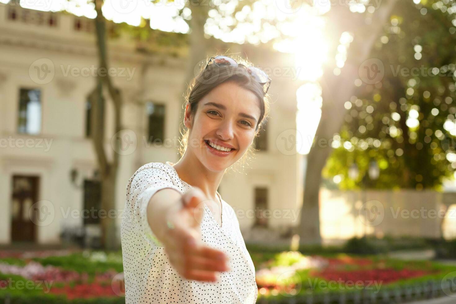 Beautiful young dark haired brunette brown-eyed female raising hand in welcome gesture while looking happily at camera and smiling broardly, wearing sunglasses on her head photo