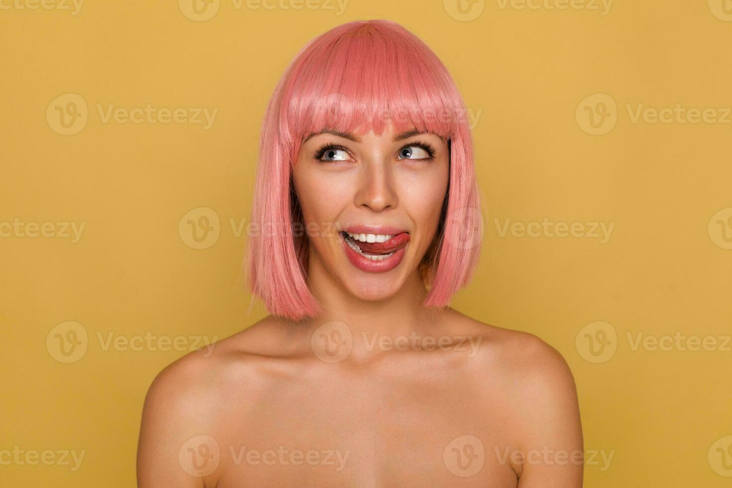 Studio shot of young lovely woman with short pink hair looking cheerfully upwards and sticking out her tongue while standing over mustard background with naked shoulders photo