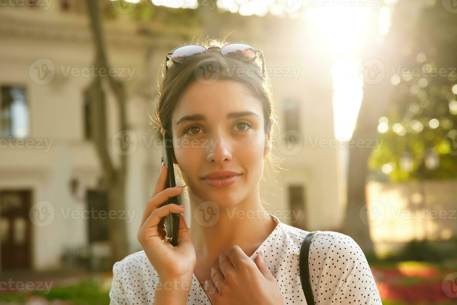 Close-up of attractive young dark haired woman with sunglasses on her head looking in front of her while talking on phone, walking over city background on sunny warm day photo