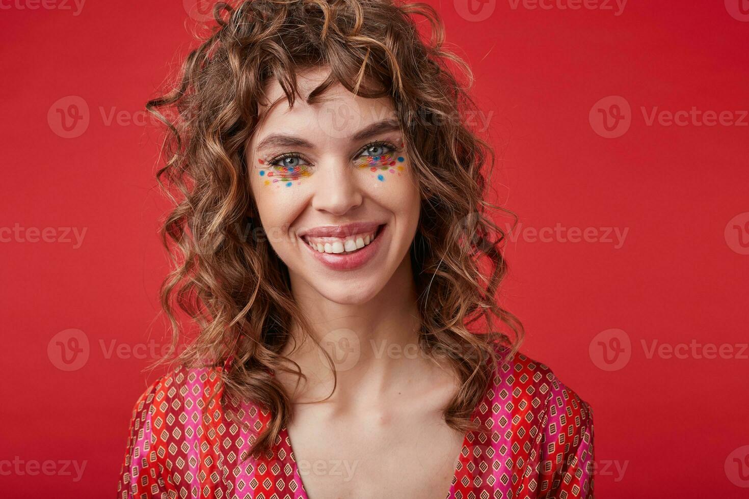 Beautiful cheerful young lady with curly hair and festive makeup standing over red background in motley patterned top, looking at camera with happy wide smile photo