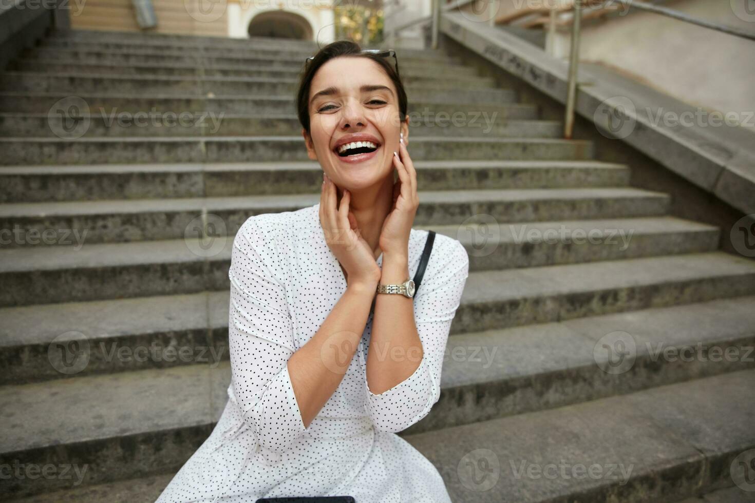 Overjoyed young attractive brunette lady with casual hairstyle holding face with raised hands and looking happily to camera, sitting over city stairs in white polka-dot dress photo