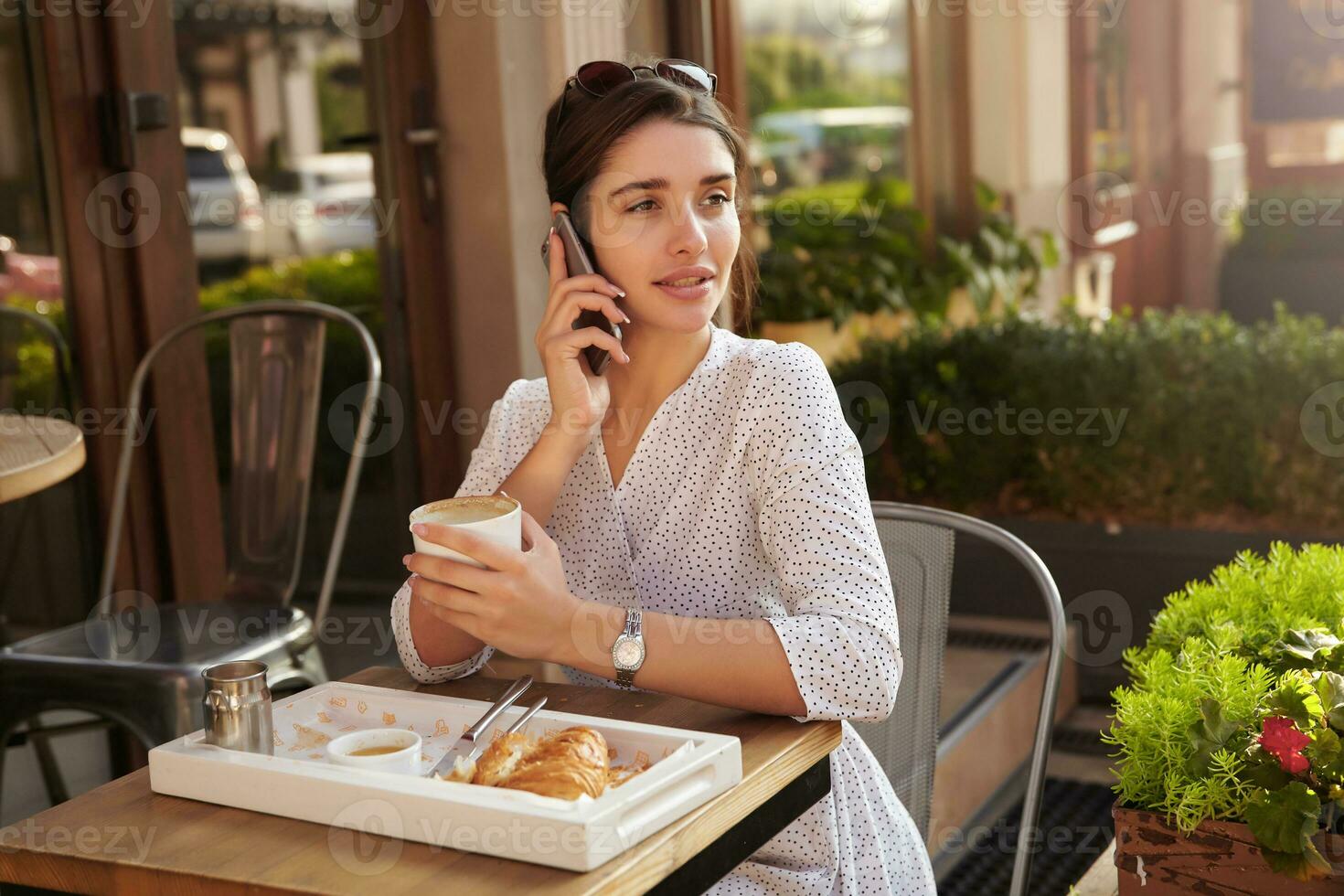 Outdoor shot of charming young dark haired lady in elegant clothes sitting at table on summer terrace and drinking coffe, looking thoughtfully aside while having phone conversation photo