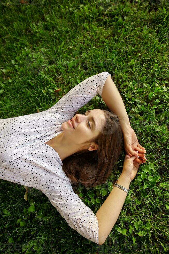Pleasant looking young attractive dark haired woman with natural makeup relaxing outdoor on sunny day with hands under her head, lying on green lawn and smiling slightly photo