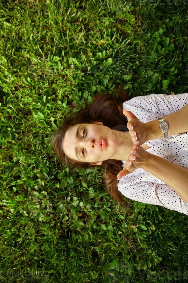 Portrait of young pretty long haired brunette female raising palms and folding lips in air kiss while looking positively at camera, posing over green grass in elegant clothes photo