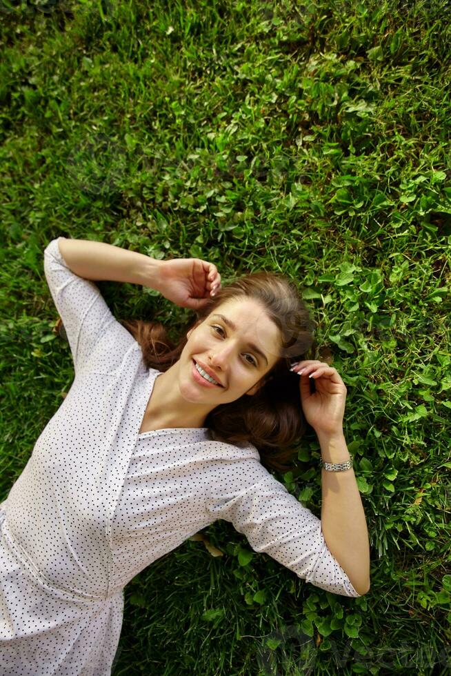 Beautiful young dark haired female in white polka-dot dress lying on green grass and raising hands to her head, being in nice mood and smiling cheerfully at camera photo