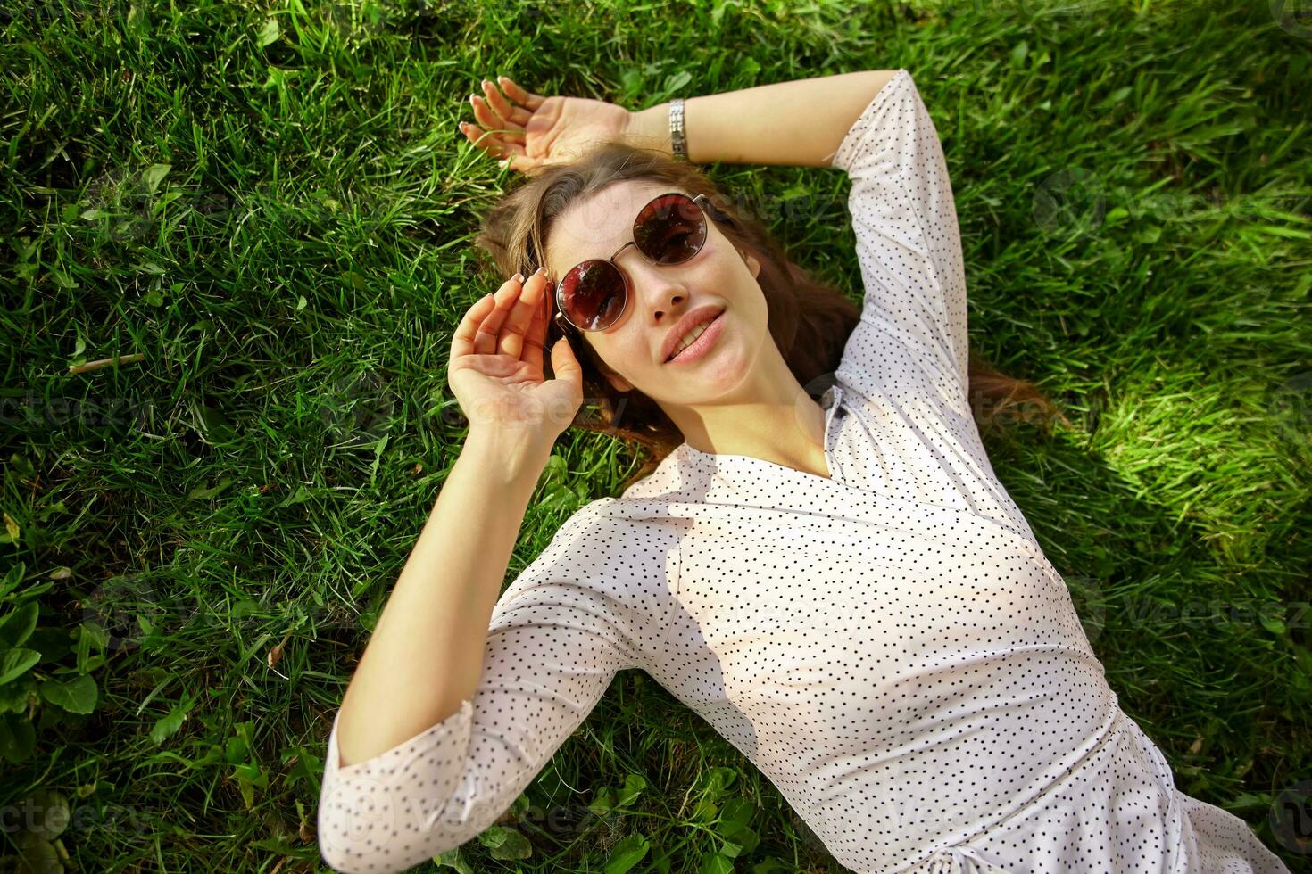 Outdoor portrait of beautiful young long haired brunette lady wearing sunglasses and white polka-dot dress while relaxing over green grass with raised hands, looking calmly at camera photo