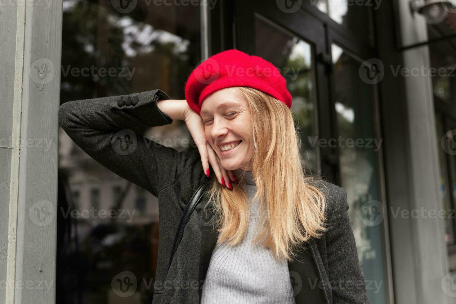 Good looking happy young blonde female with casual hairstyle keeping her eyes closed while smiling pleasantly, leaning on windowsill while standing over cafe exterior photo