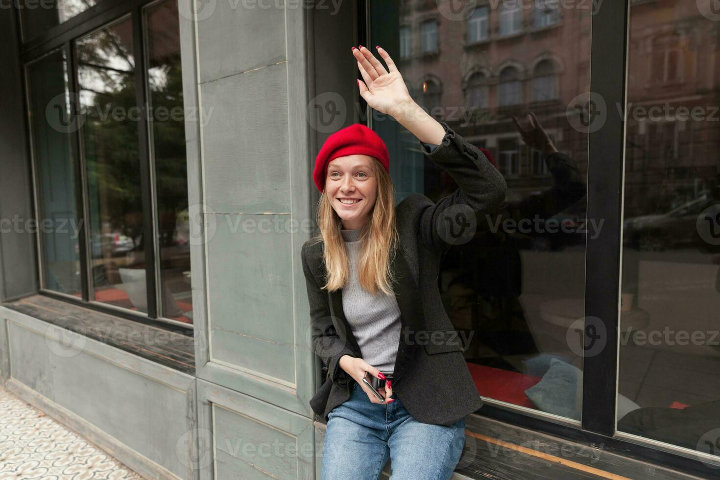 al aire libre Disparo de atractivo joven alegre dama vistiendo su largo rubia pelo en casual peinado, vestido en de moda ropa mientras reunión su amigos en fin de semana, levantamiento palma en Hola gesto foto