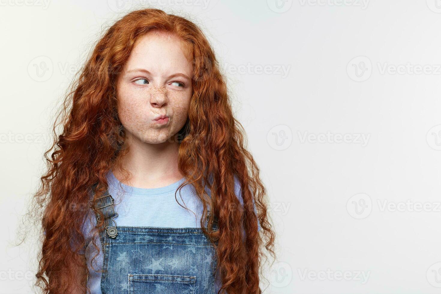 Portrait of displeased cute freckles little girl with ginger hair, looks with disgust at the copy space on the left side, tands over white background. photo