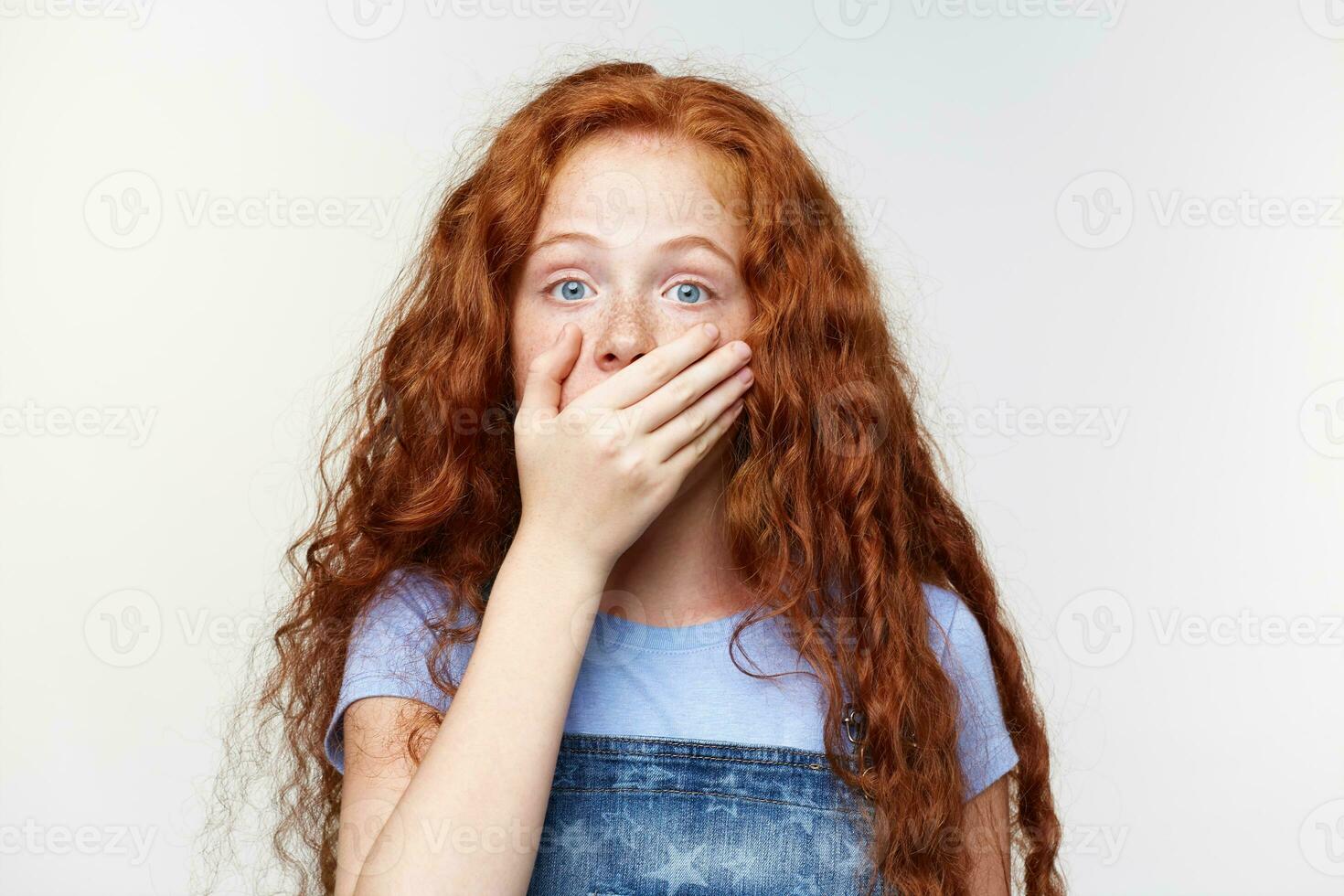 Portrait of shocked cute freckles little girl with ginger hair, hears unbelievable news , covered mouth with hand, stands over white wall with wide open eyes with surprised expression. photo