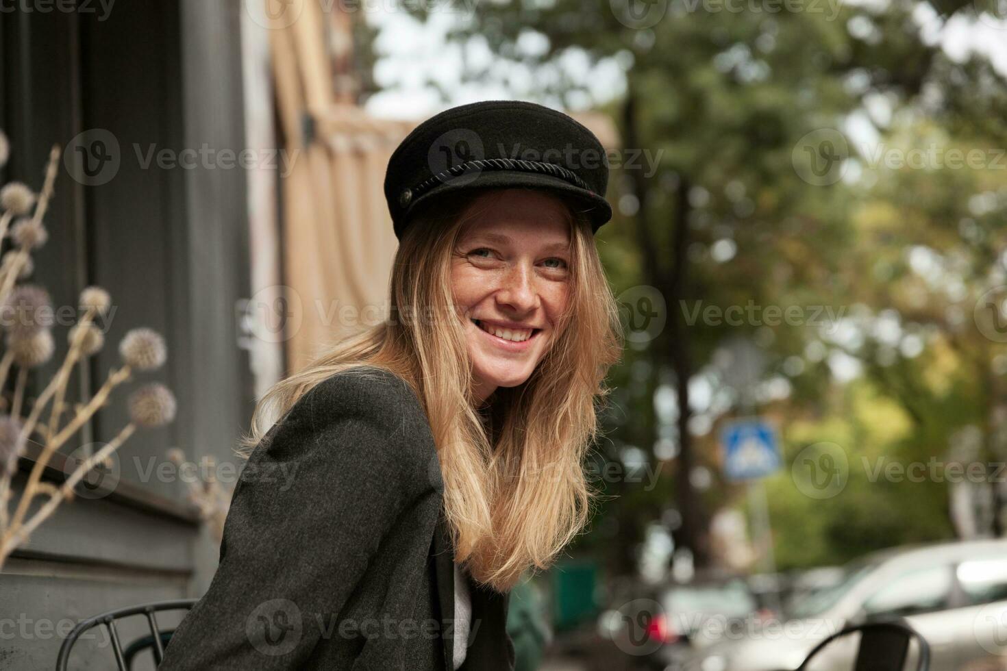 Portrait of young pleasant looking female with long blonde hair posing over city background on sunny weekend, being in nice mood and smiling sincerely photo