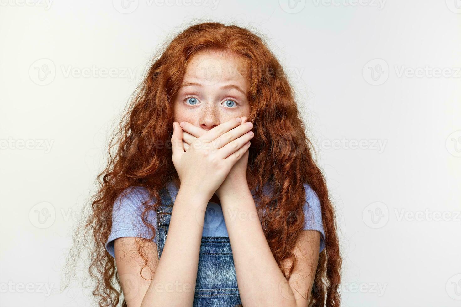 Portrait of scared cute freckles little girl with ginger hair, hears heard a terrible storry, covered mouth with palms, stands over white wall with wide open eyes with afraided expression. photo