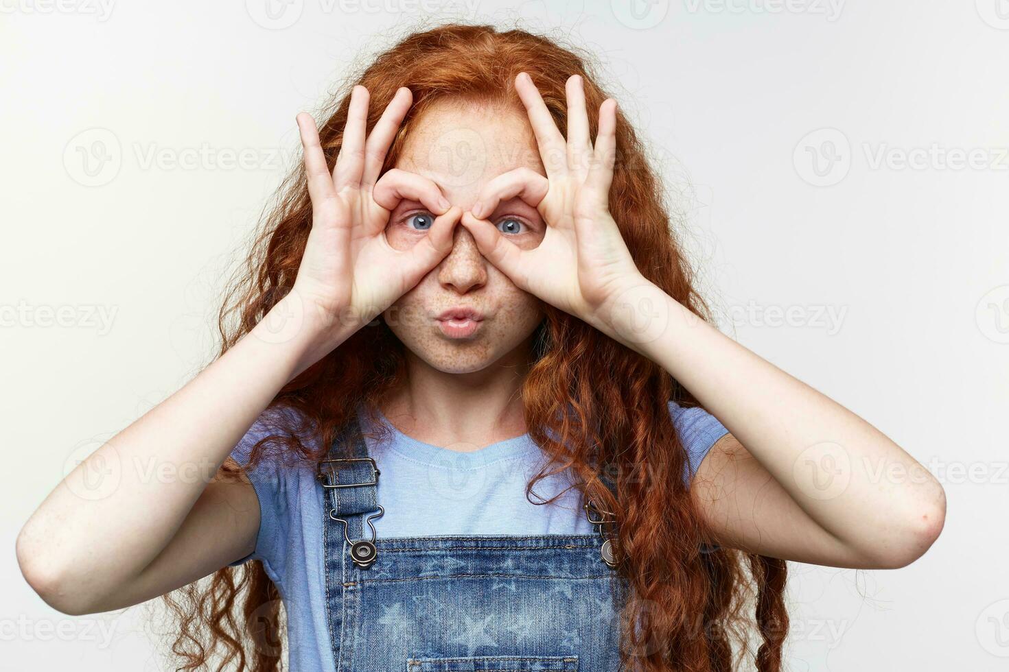 cerca arriba de gracioso alegre bonito pequeño niña con jengibre pelo y pecas, mira mediante manos, mira lindo, soportes terminado blanco antecedentes. foto