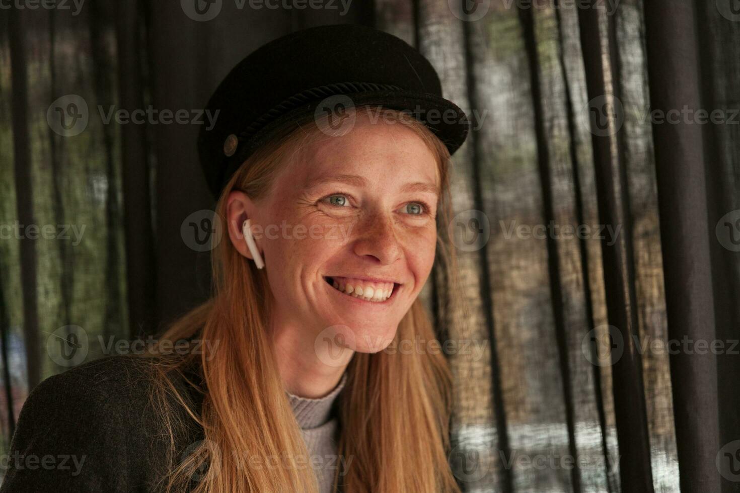 Close-up of happy blue-eyed young blonde lady with natural makeup wearing earphones and looking cheerfully ahead with broad sincere smile, posing over cafe interior photo
