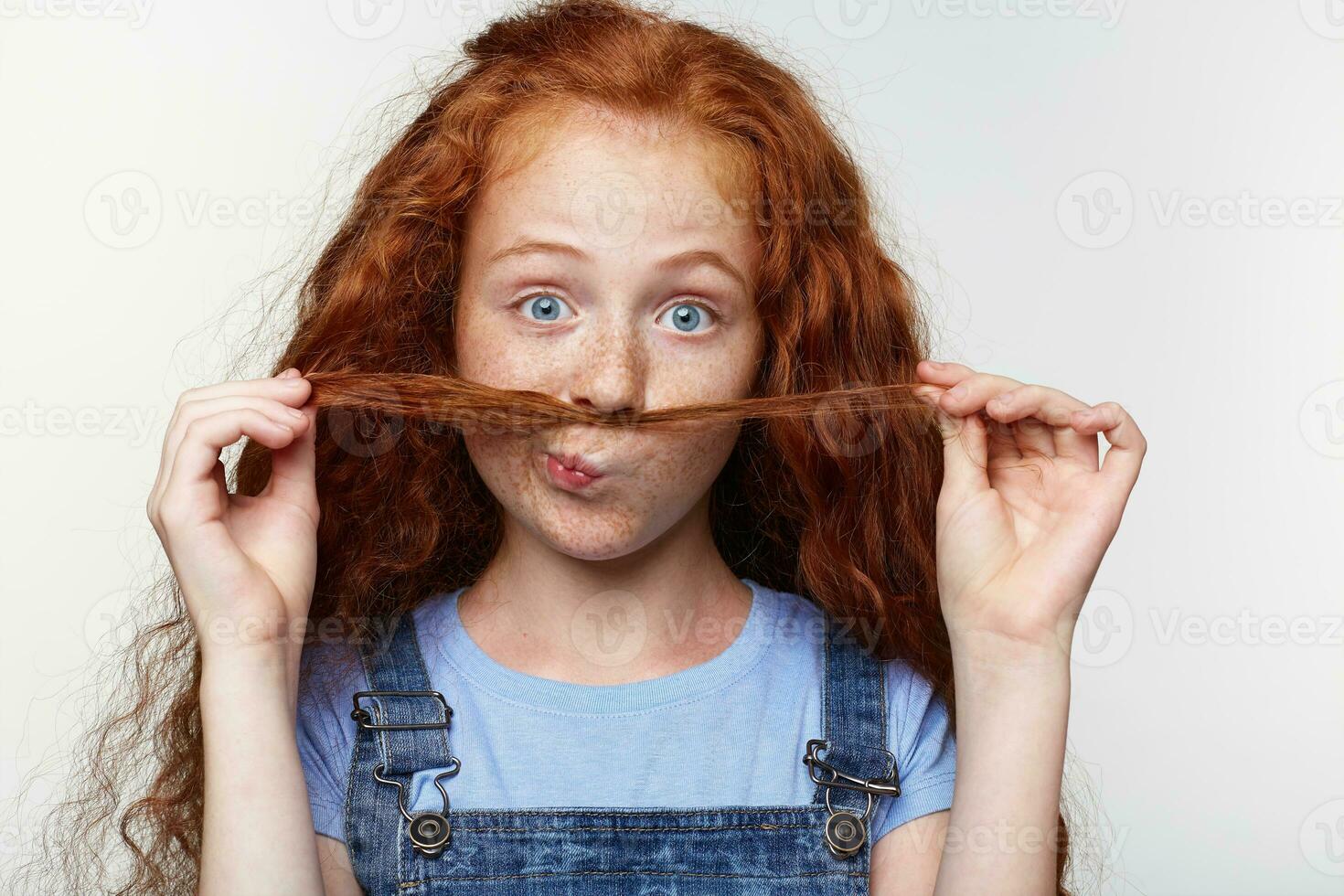 Close up of joyful cute little girl with ginger hair and freckles, looks at the camera, makes a mustache from strands of hair and looks funny, stands over white background. photo