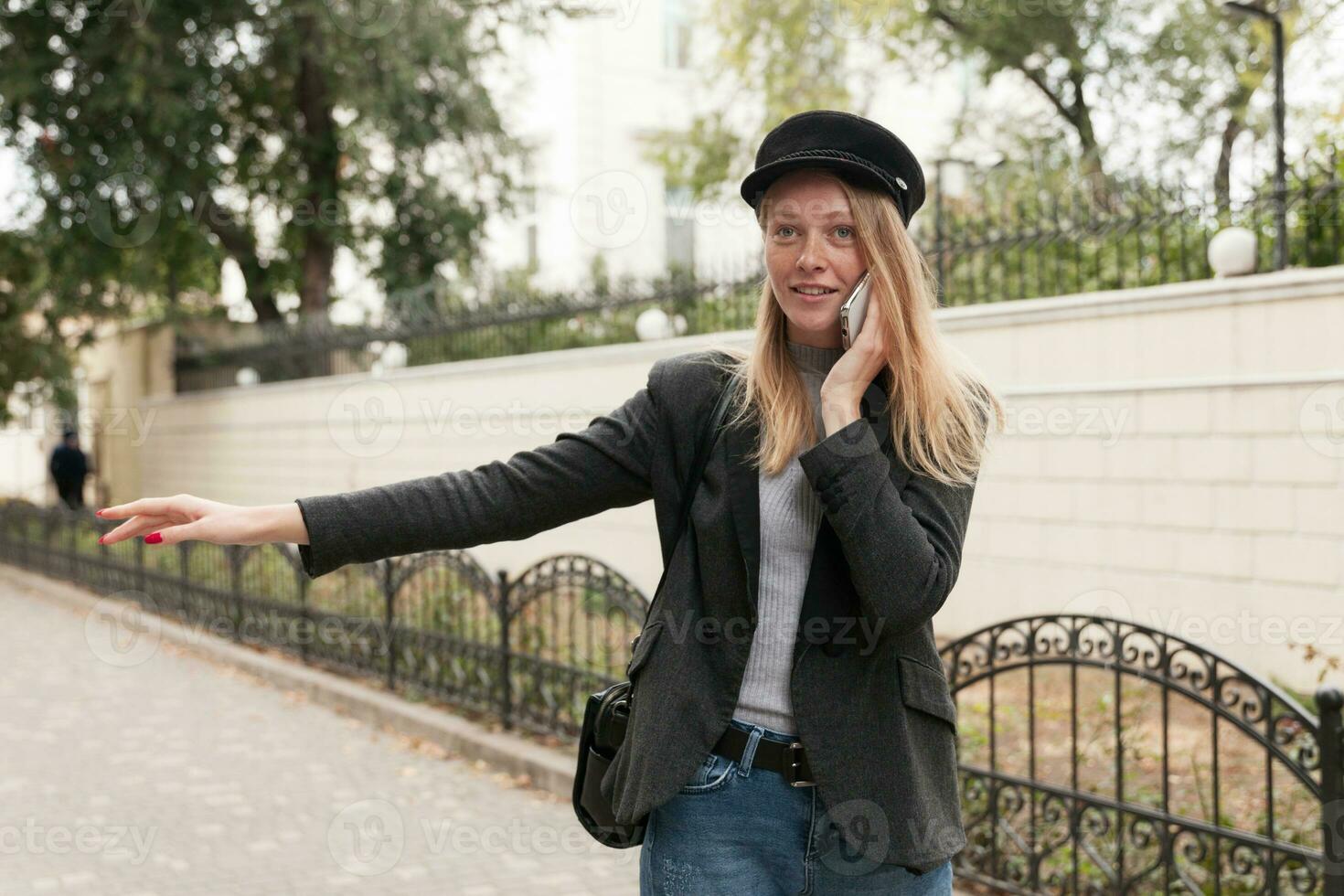 Outdoor photo of positive young pretty blonde female dressed in trendy clothes while standing over city background, catching taxi with raised hand while making call with her smartphone