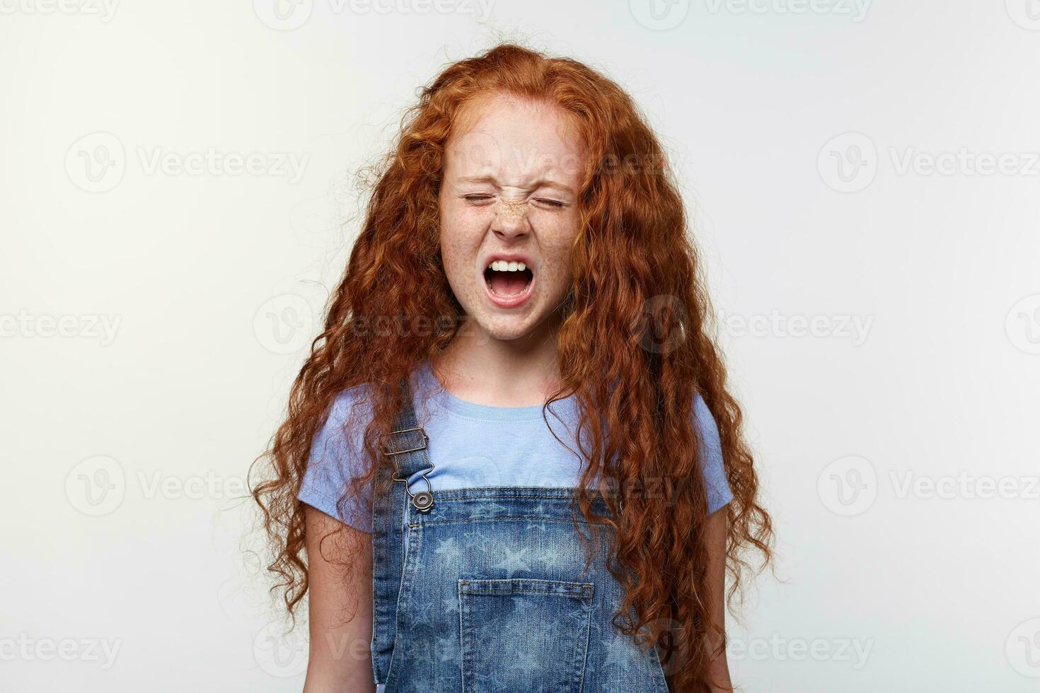 Prtrait of sceaming freckles little girl with ginger hair, stands over white wall with closed eyes and wide open mouth, looks unhappy and crying. photo