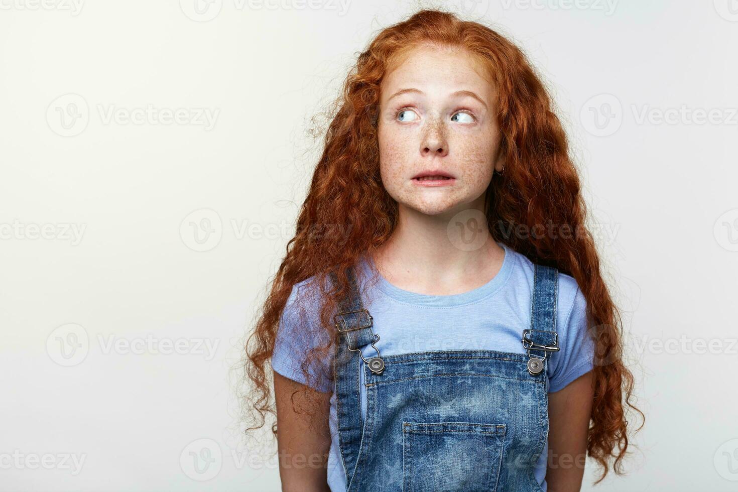 Portrait of guilty cute freckles little girl with ginger hair, pretends not to do anything wrong, looks away over white background with copy space on the left side. photo
