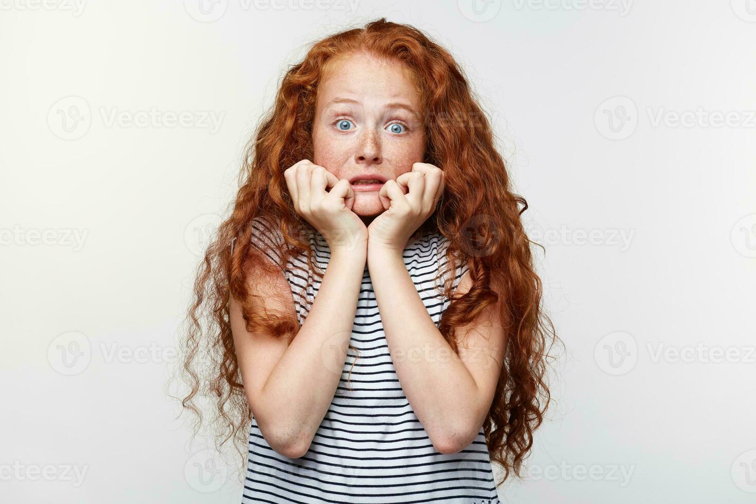 Portrait of afraid little girl with ginger hair and freckles, scared and anxious biting her finger nails, looking at camera with wide opened eyes and looks away, isolated over white wall. photo