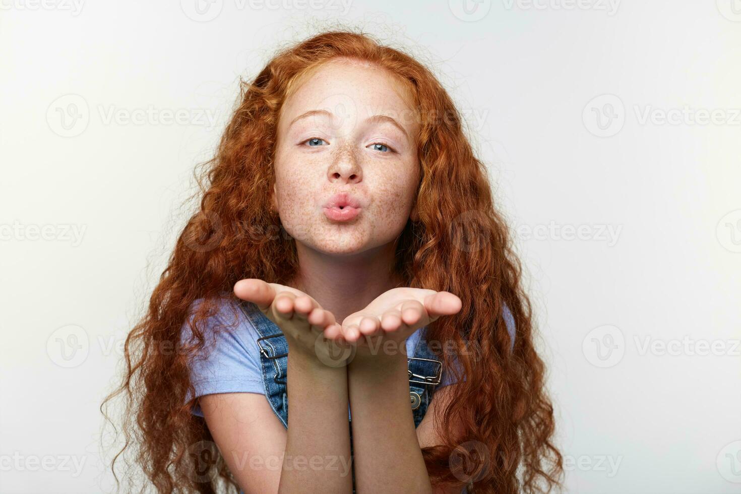 Portrait of cheerful little girl with ginger hair and freckles, send kiss at the cam, looks happy, standing over white wall. photo