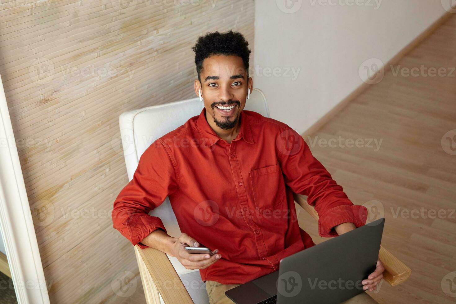 Cheerful young brown-eyed brunette bearded dark skinned man looking gladly at camera with charming smile while working out of office with his mobile phone and laptop photo