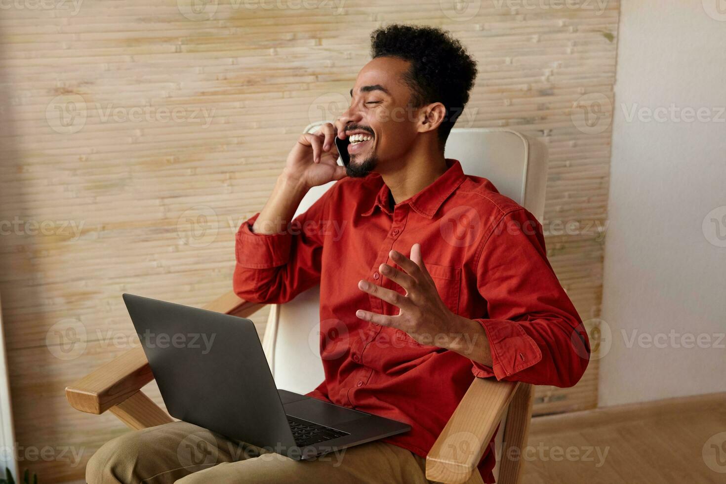 Side view of good looking young cheerful short haired bearded male with dark skin keeping his eyes closed while having pleasant talk, sitting in chair over home interior photo