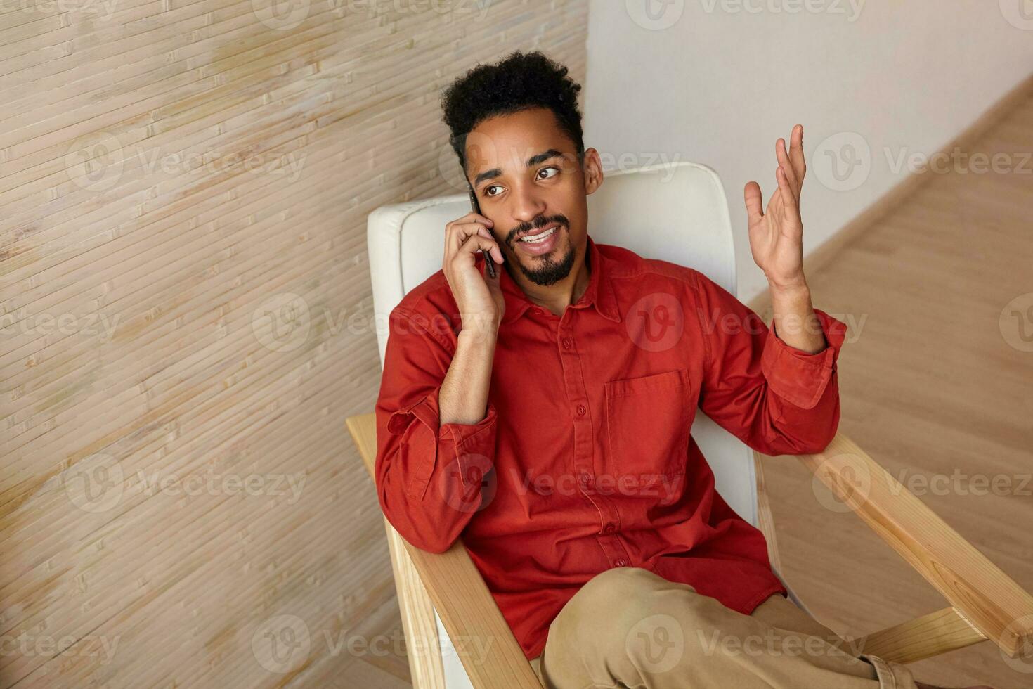 Glad young pretty bearded brunette dark skinned male with short haircut keeping his hand raised while having phone talk and looking positively aside, isolated over home interior photo