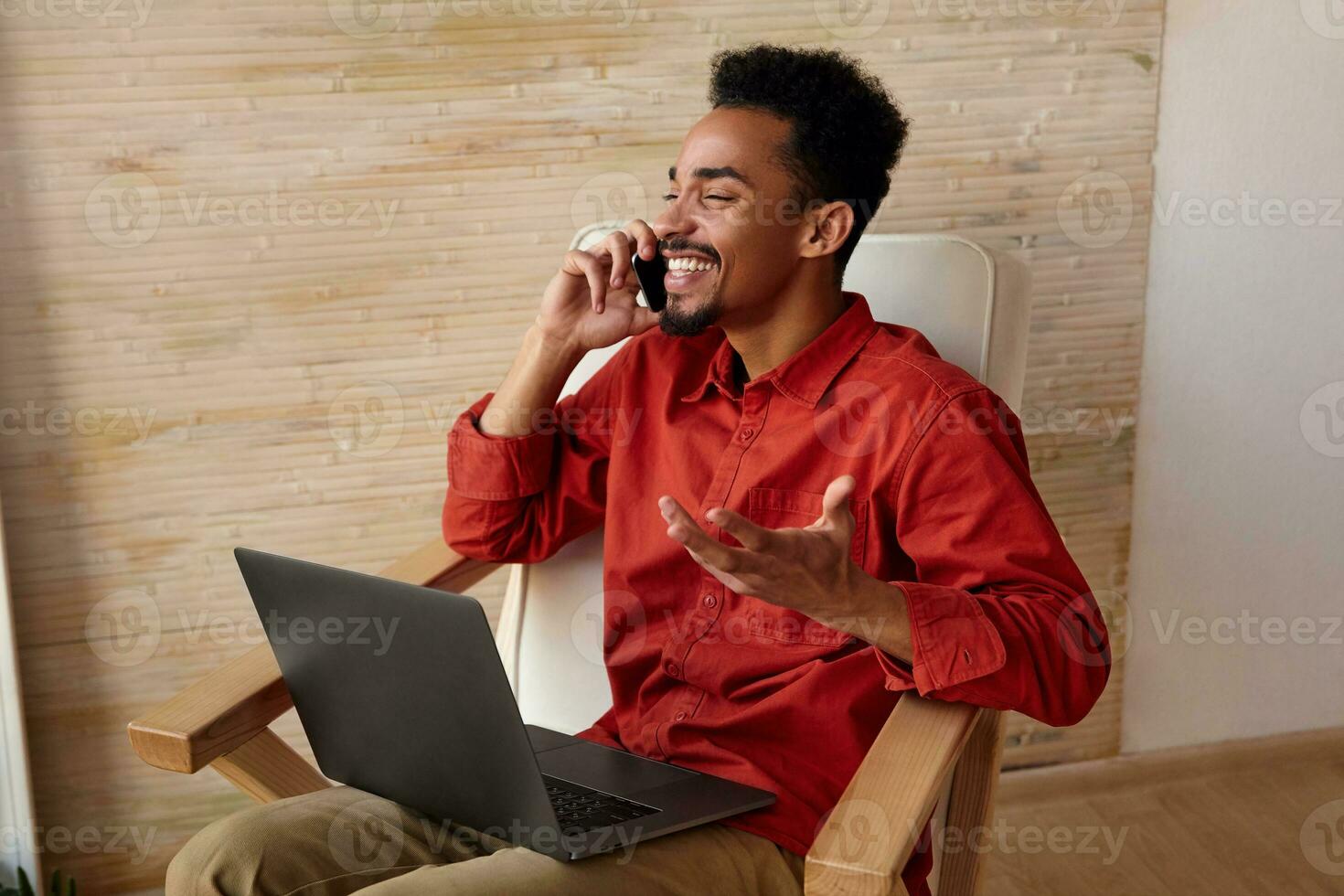 Indoor photo of young joyful bearded dark skinned male with short haircut laughing happily with closed eyes while having phone talk, isolated over beige interior