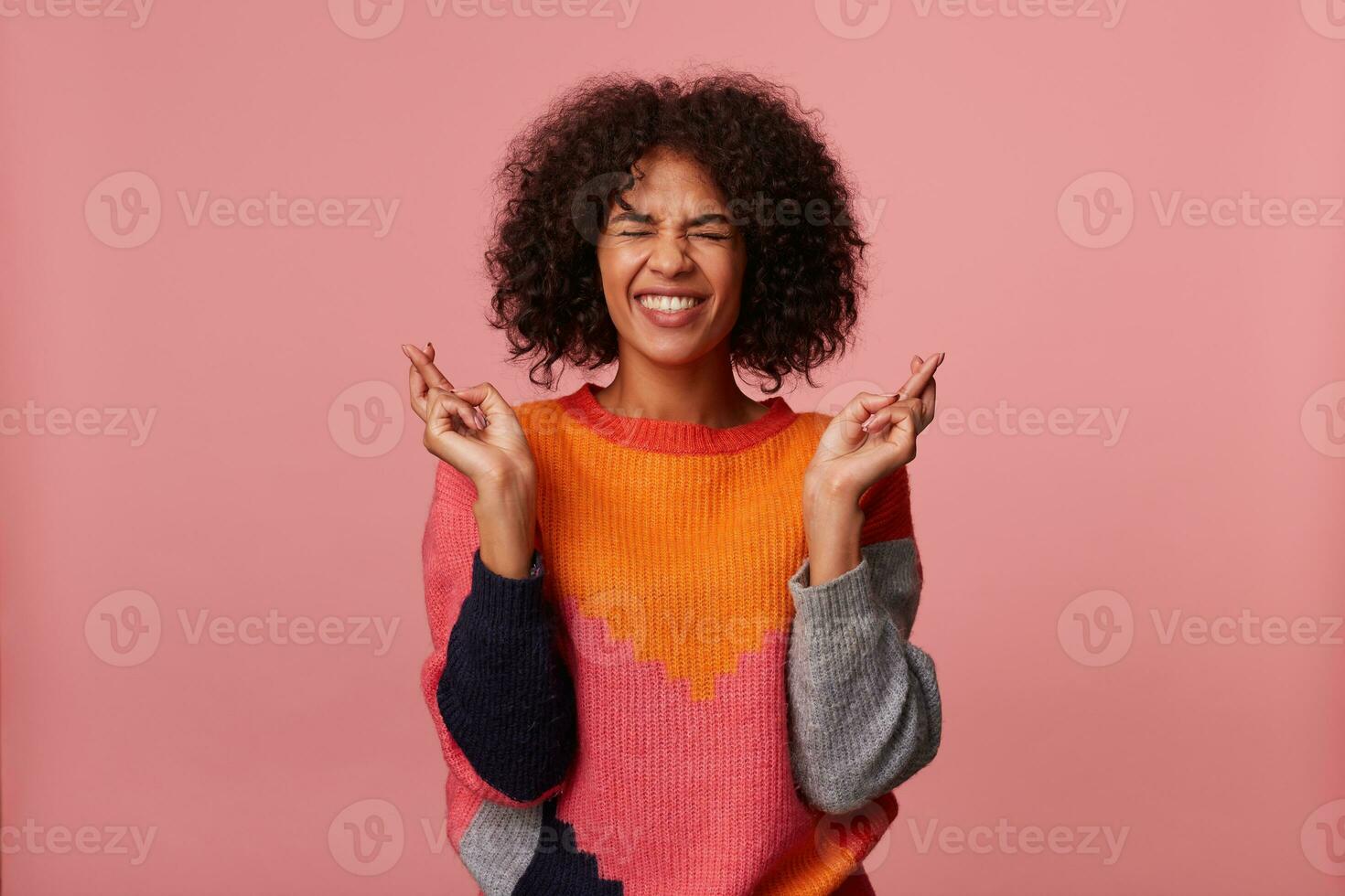 Pleasant looking young joyful dark skinned lady with short curly brown hair raising hands with crossed fingers, keeping eyes closed while posing over pink background in casual wear photo