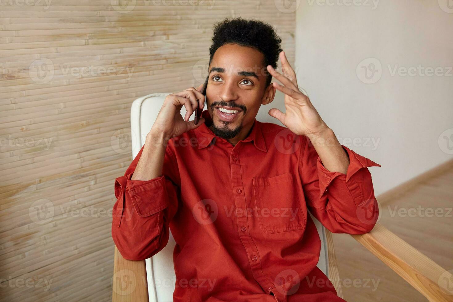 Positive young lovely dark haired bearded male dressed in red shirt leaning his head on raised hand while having pleasant phone conversation, isolated over home interior photo