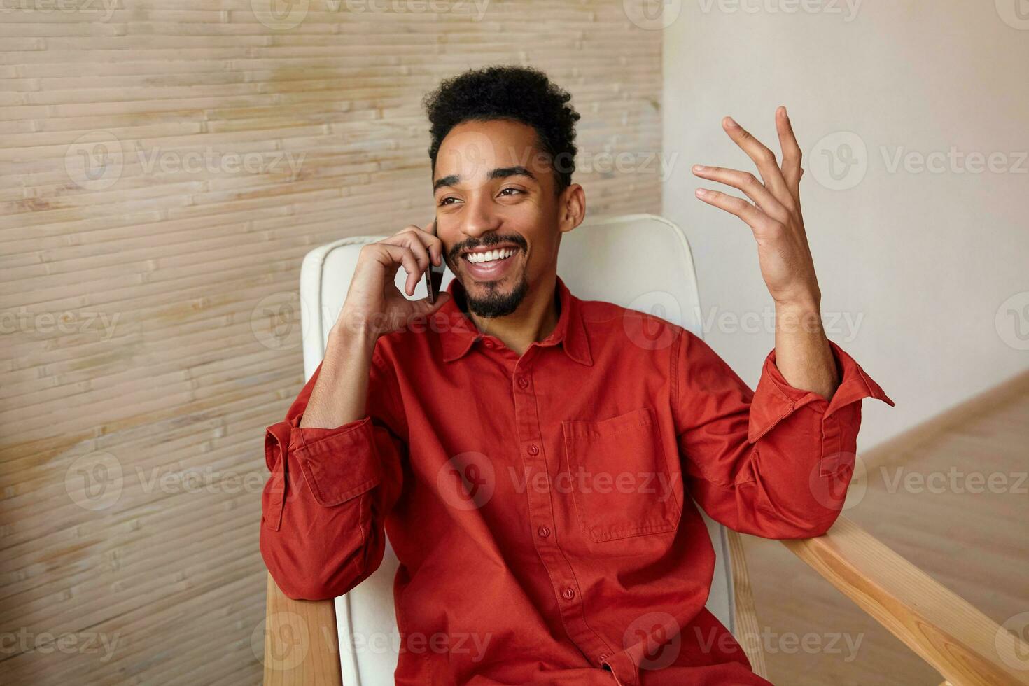 Horizontal shot of young lovely glad dark skinned male with short haircut smiling cheerfully while having pone talk and raising emotionally his hand, sitting in chair over home interior photo