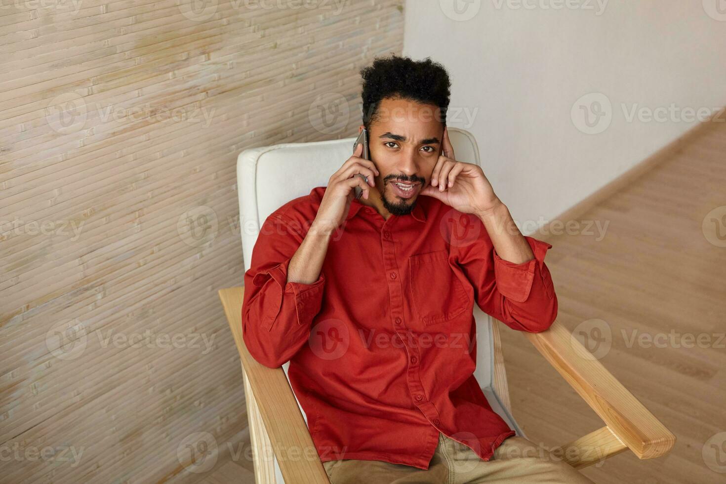 Indoor shot of young brown-eyed short haired bearded male with dark skin leaning his face on raised hand and looking confusedly at camera while making call photo