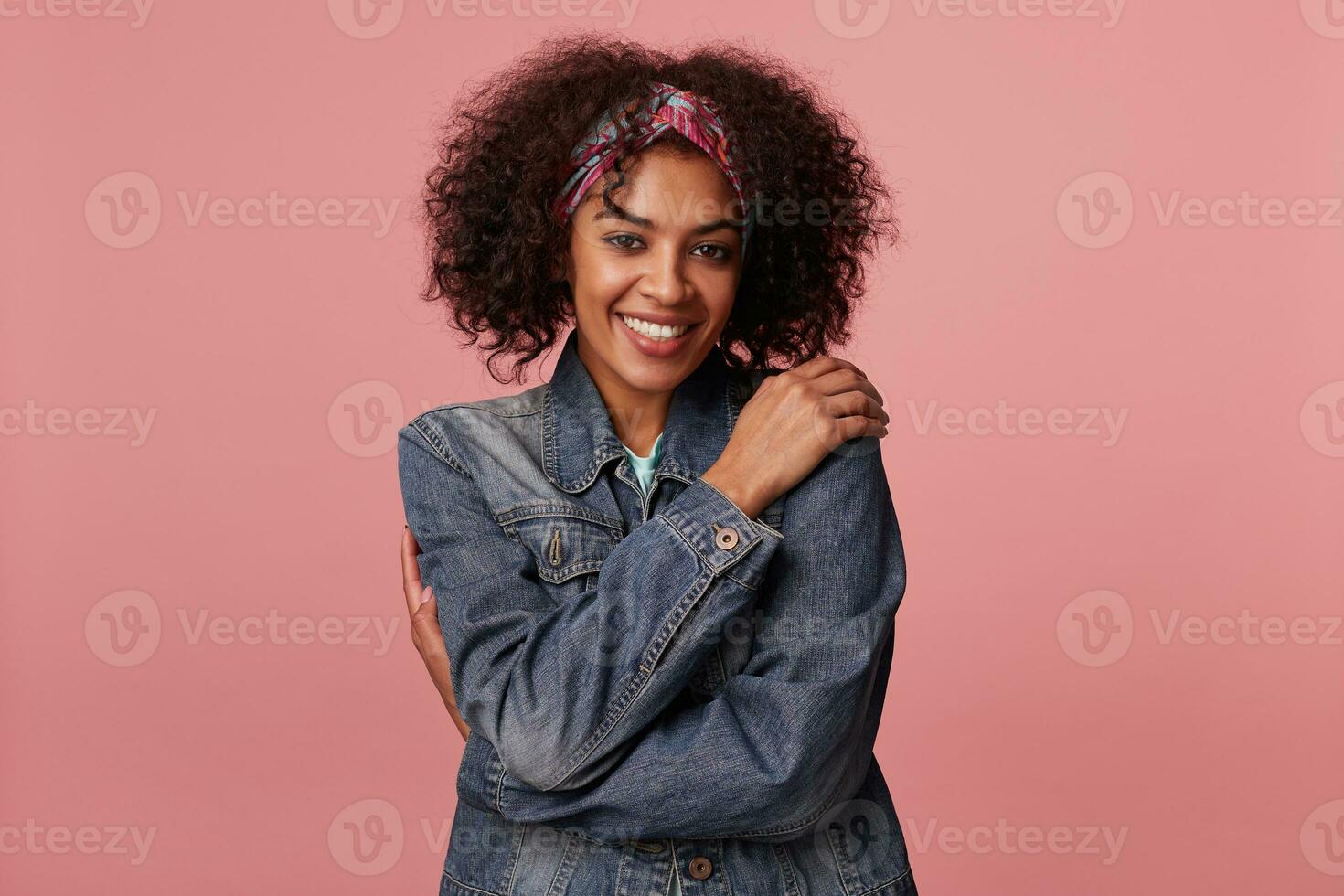 Positive attractive young dark skinned brunette lady with casual hairstyle hugging herself and looking happily to camera with charming smile, standing against pink background photo
