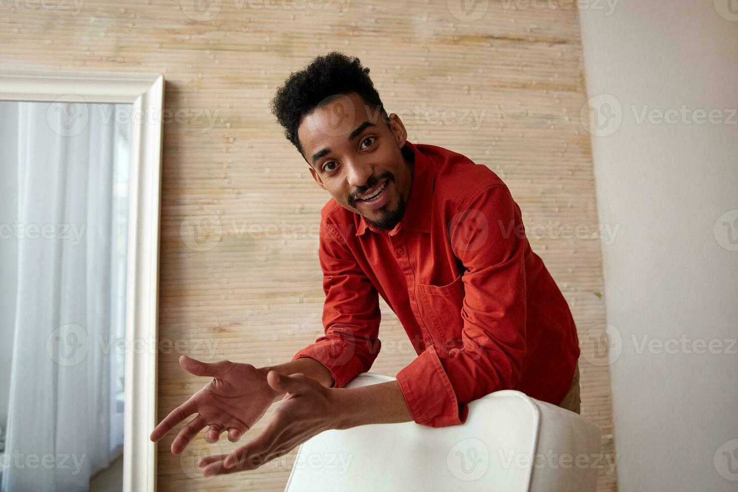 Glad young brown-eyed dark skinned brunette guy with short haircut looking positively at camera while leaning on chair, standing over home interior in red shirt photo