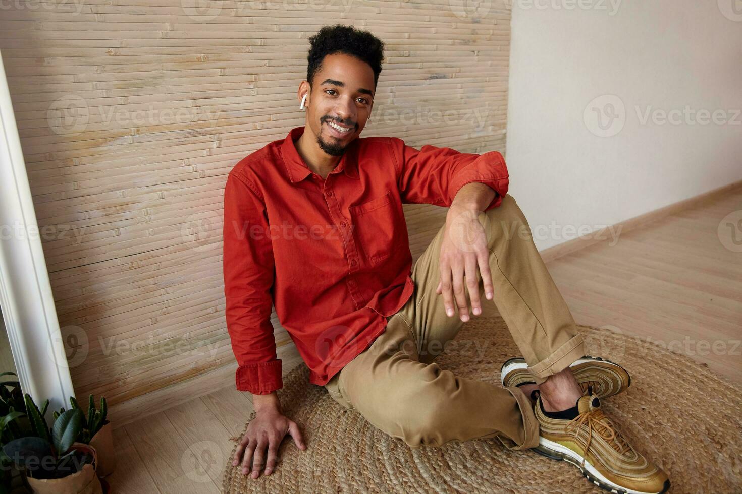 Happy young handsome short haired curly dark skinned male dressed in casual wear crossing his legs while sitting on floor and looking joyfully at camera, isolated over beige interior photo