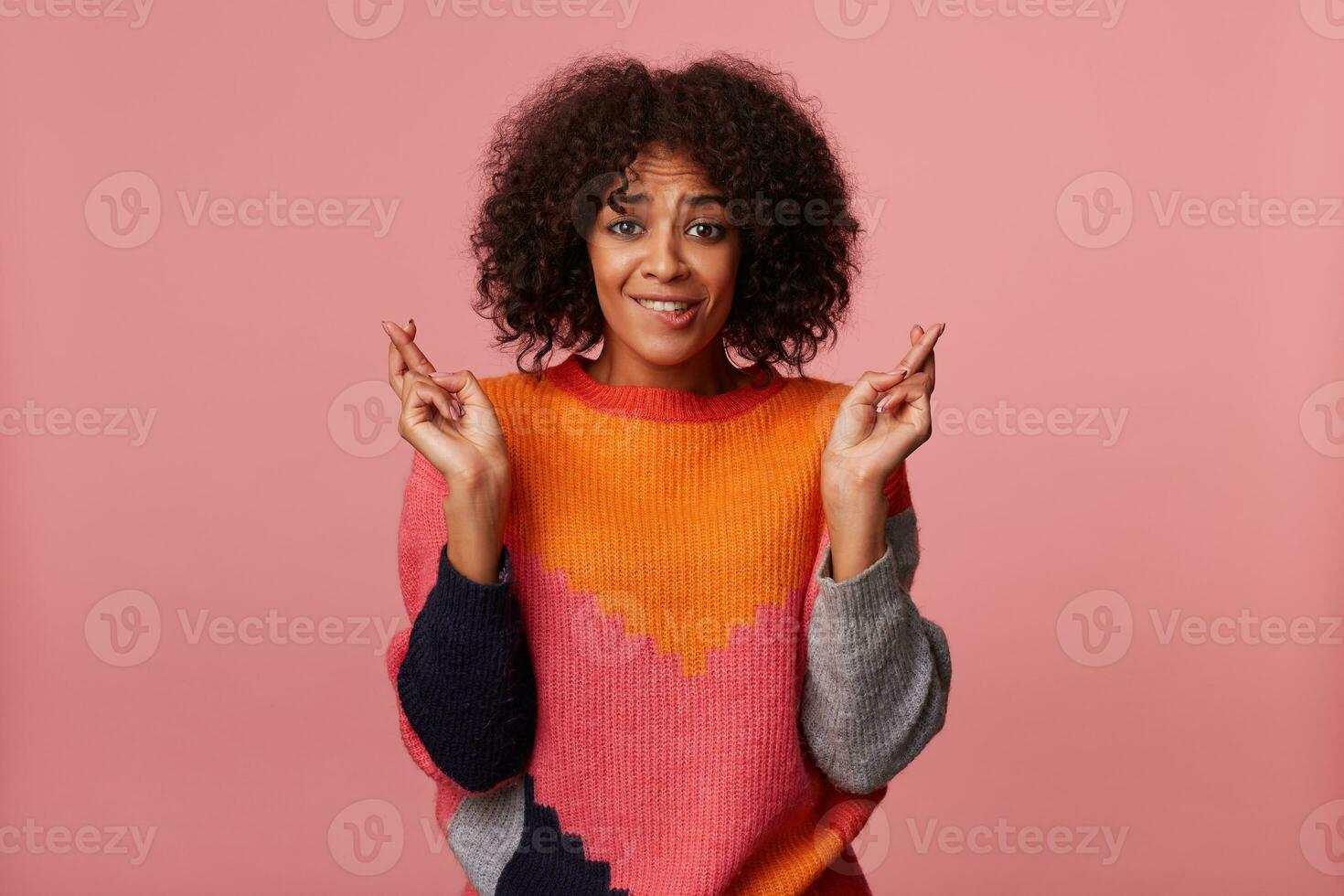Portrait of young pretty curly brunette female with dark skin wearing casual hairstyle while standing over pink background, crossing fingers for good luck and biting underlip photo