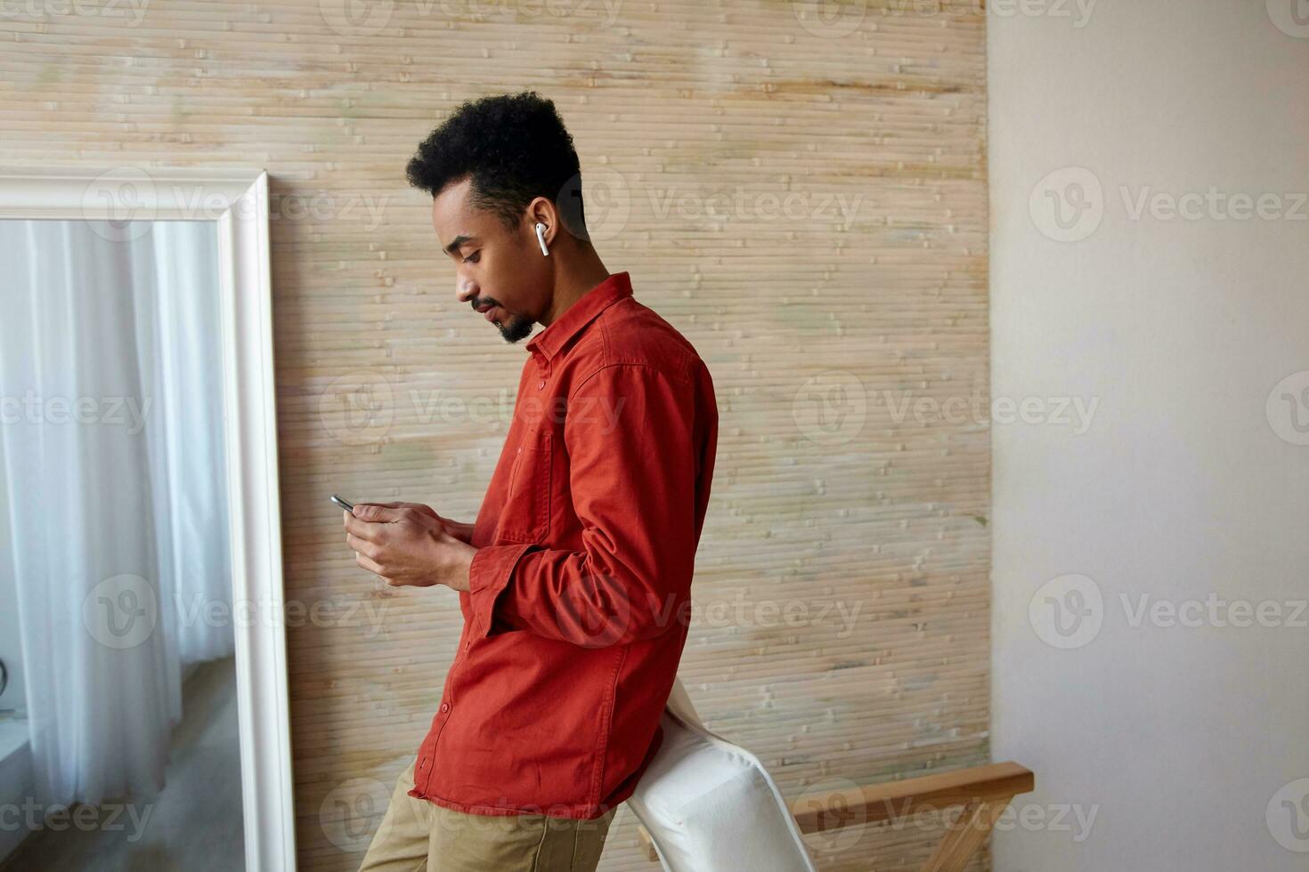 Side view of attractive young dark skinned brunette man with beard leaning on chair while checking his social networks, standing over home interior in casual wear photo